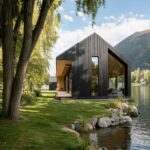 Riverstone Retreat, a charred timber gabled home in Poland set among trees at the water's edge with mountain views